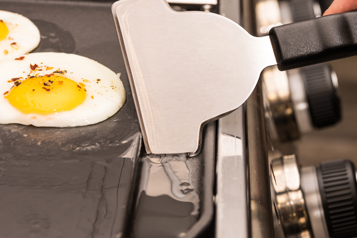 Scraper clearing the edge of a griddle beside a fried egg
