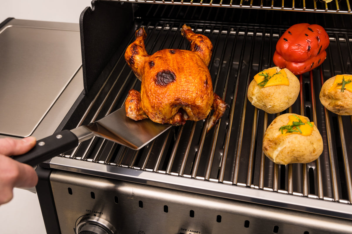 Wide stainless steel turner lifting roast chicken from a grill