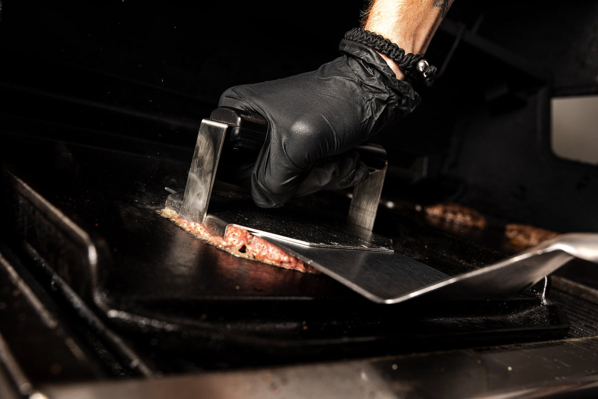 Gloved hand using a smash burger press on a separate cooking surface