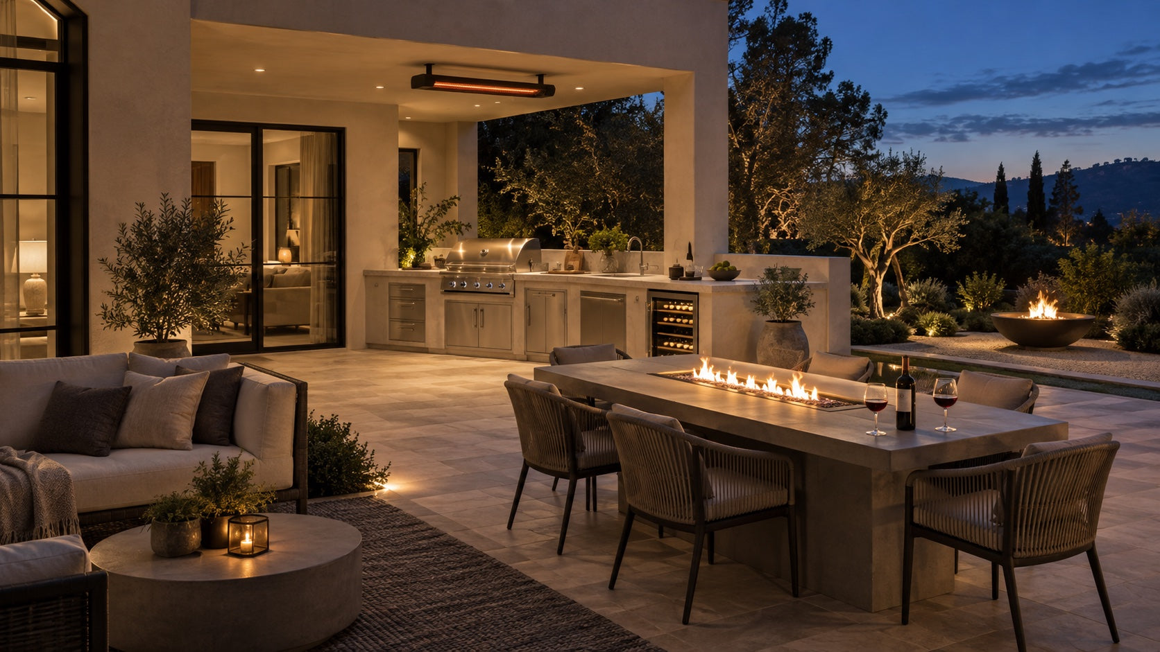 An outdoor gathering space with a kitchen, dining table and comfortable lounge seating at dusk